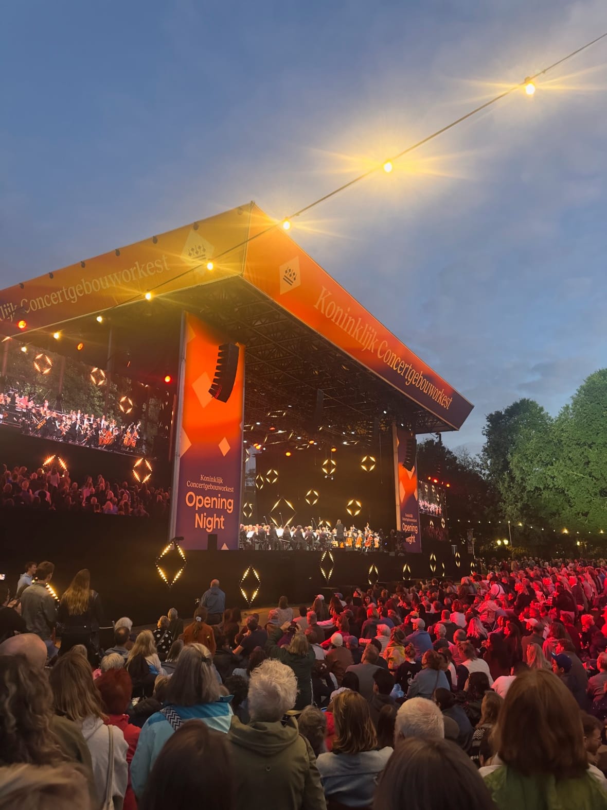 Bezoekers op het gras van het Oosterpark bij schemering, met het verlichte podium op de achtergrond