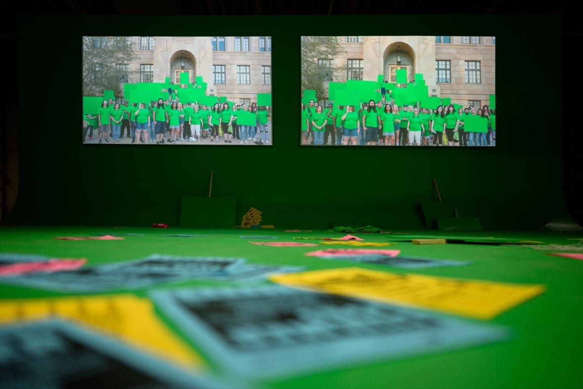 Installatiebeeld bij Framer Framed: twee schermen met een groep mensen in groene T-shirts met groene protestborden, daaronder posters op de vloer