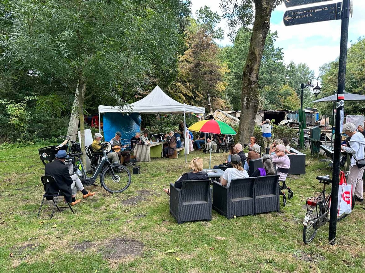 Bezoekers luisteren naar live muziek op het terrein van Parknest, met de uitgebrande resten op de achtergrond