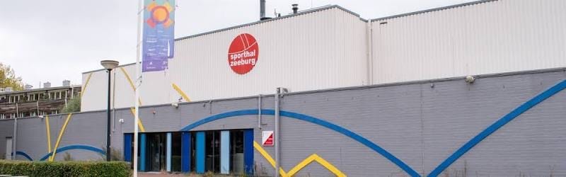 Het gebouw van Sporthal Zeeburg met de blauw-gele golflijn op de grijze gevel en de glazen ingang